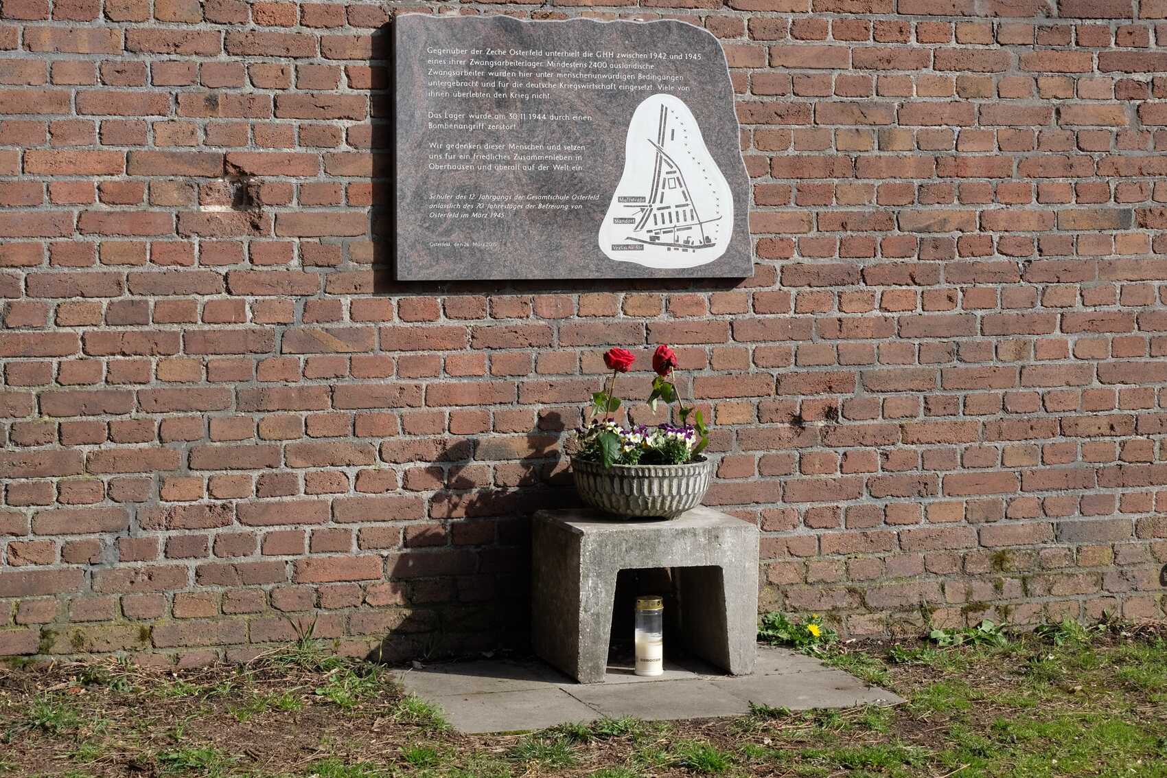 Informationstafel aus Stein mit Hinweise auf das Zwangsarbeiterlager der Gutehoffnugshütte, davor Hocker aus Stein mit Blumenschale