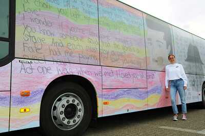 A schoolgirl is standing at the side of a brightly printed STOAG bus